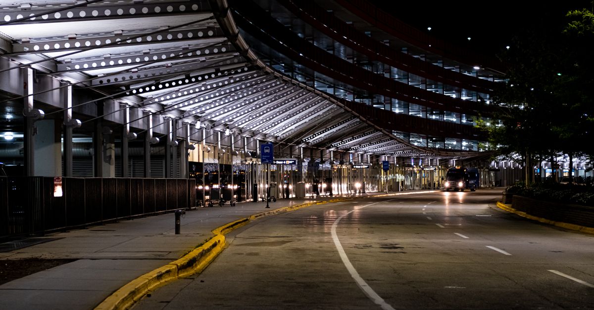 An empty airport terminal kerbside and canopy lit up at night, with a shuttle bus approaching.