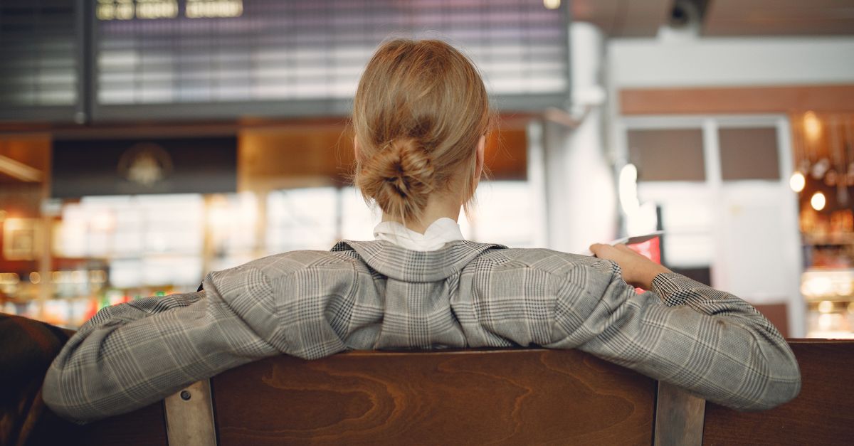 A traveller sitting in an airport terminal, holding a boarding pass, with a blurred departures board behind