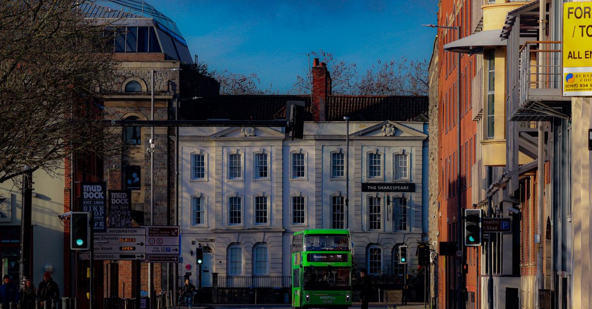 A green double-decker bus passing Georgian buildings on a Bristol street, with pub and shop signs including Mud Dock and The Shakespeare